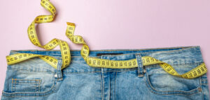Image of the waist of jeans and yellow measuring tape instead of belt on pink background.