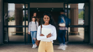 Portrait of Black girl student standing at university campus with other students walking in background. Young woman standing in college with students walking at the back.