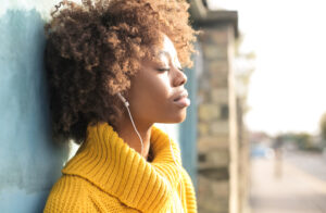 Image of a Black female presenting person closing her eyes, listening to music outside. Calm. 