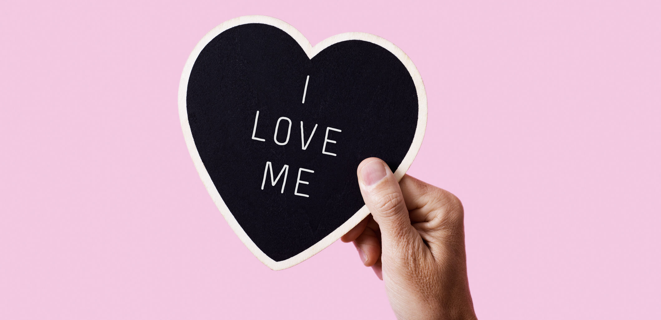 closeup of a man holding a black heart-shaped sign with the text I love me written in it, on a pink background
