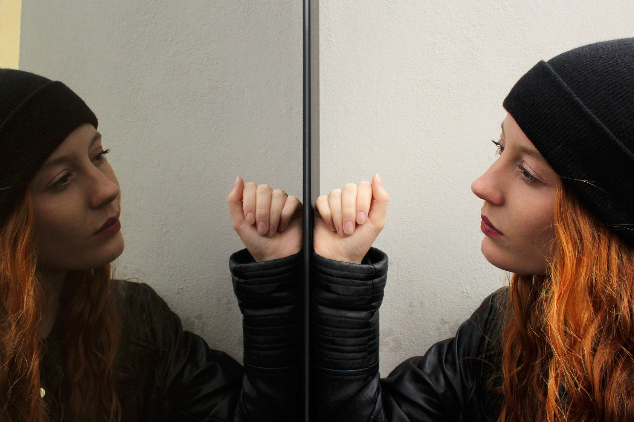 Girl with long red hair wearing a black woolen cap and a black leather jacket looking at herself mirrored on a glass door with a thoughtful expression. Horizontal