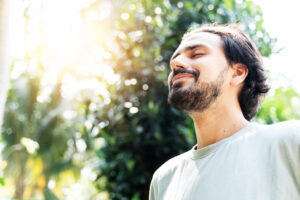 Image of a bearded man is meditating outdoor in the park with face raised up to sky and eyes closed on sunny summer day