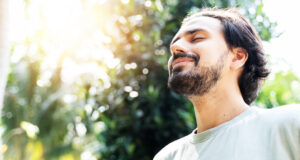 Image of a bearded man, outside with sun on his face, eyes closed, smiling.