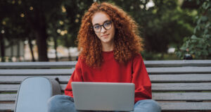Confident smiling millennial female student with red curly hair wearing casual clothes and eyeglasses using laptop while sitting on bench in city park