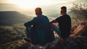 A mentor and mentee sitting on a mountain next to each other, overlooking the valley. Mentoring relationship benefiting their mental health.