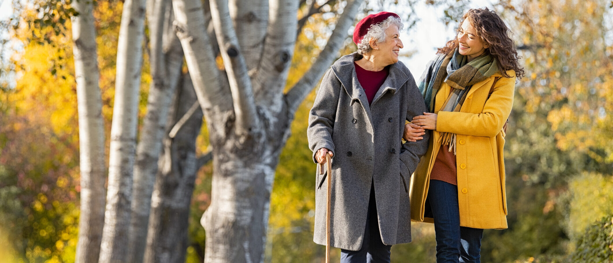 Granddaughter walking with senior woman in park wearing winter clothing. Old grandmother with walking cane walking with lovely caregiver girl in sunny day. Happy woman and smiling grandma walking in autumn park.
