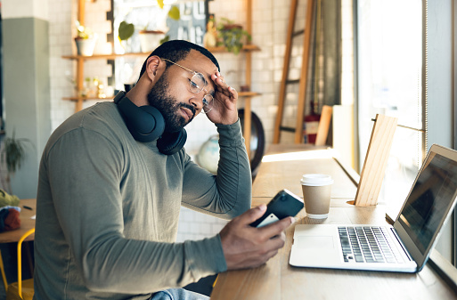 Man looking at phone in front of laptop feeling stressed and overwhelmed by news consumption and the impact on his mental health