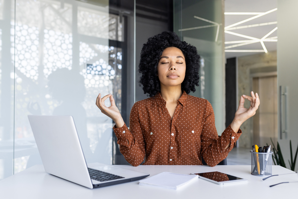 A woman sitting at her laptop, taking a break with eyes closed, practicing a mindful meditation to improve her mental health