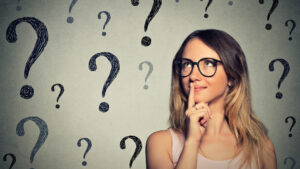 Image of a woman thinking with glasses on, finger on mouth and a wall with question marks behind her.