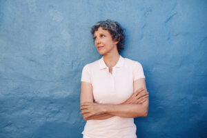 Middle-aged woman feeling calm after Brainspotting therapy. Arms crossed leaning against aa blue wall in a white shirt.