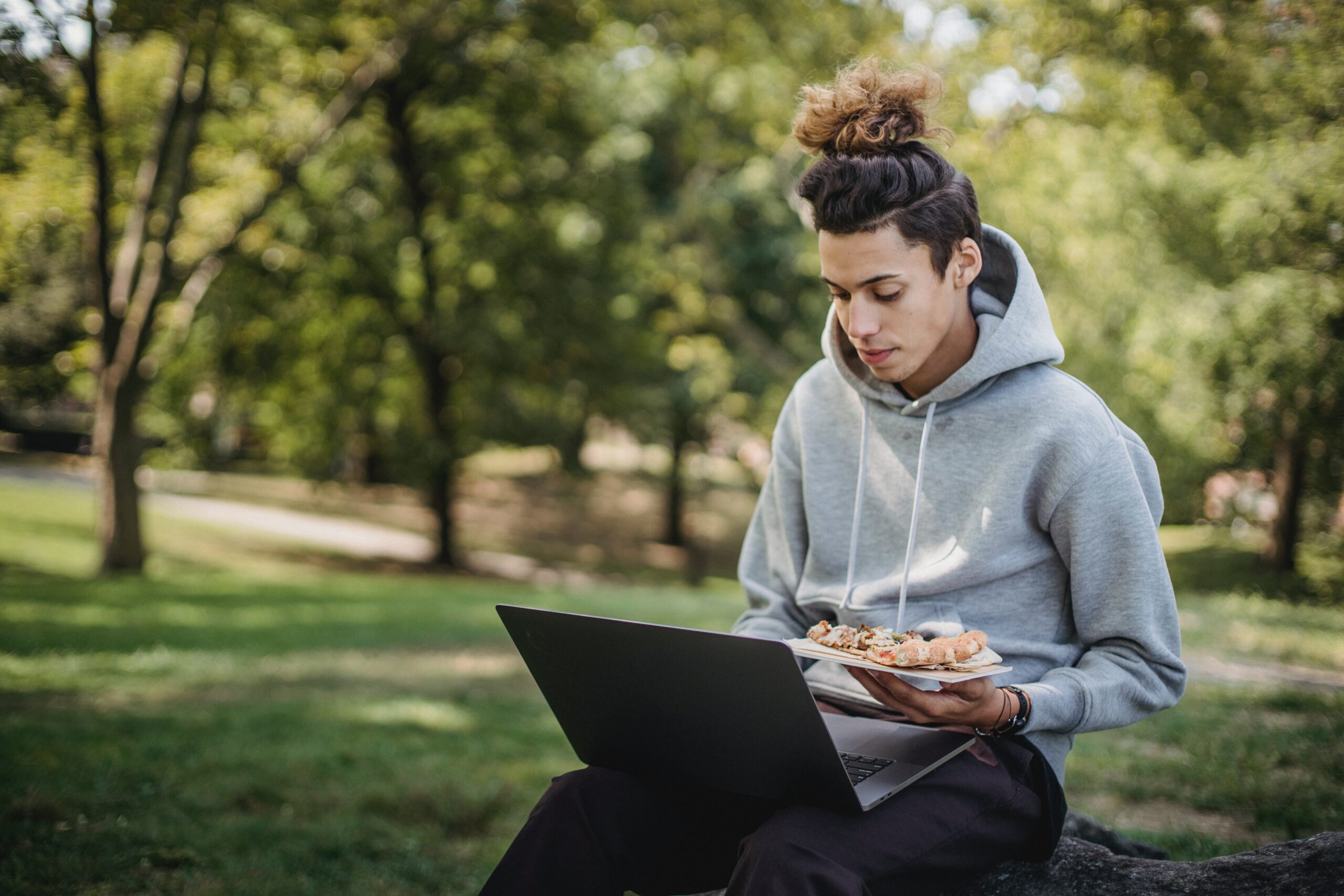 Male college student sitting on the campus lawn on his laptop holding a slice of pizza wearing a hoodie with a man bun