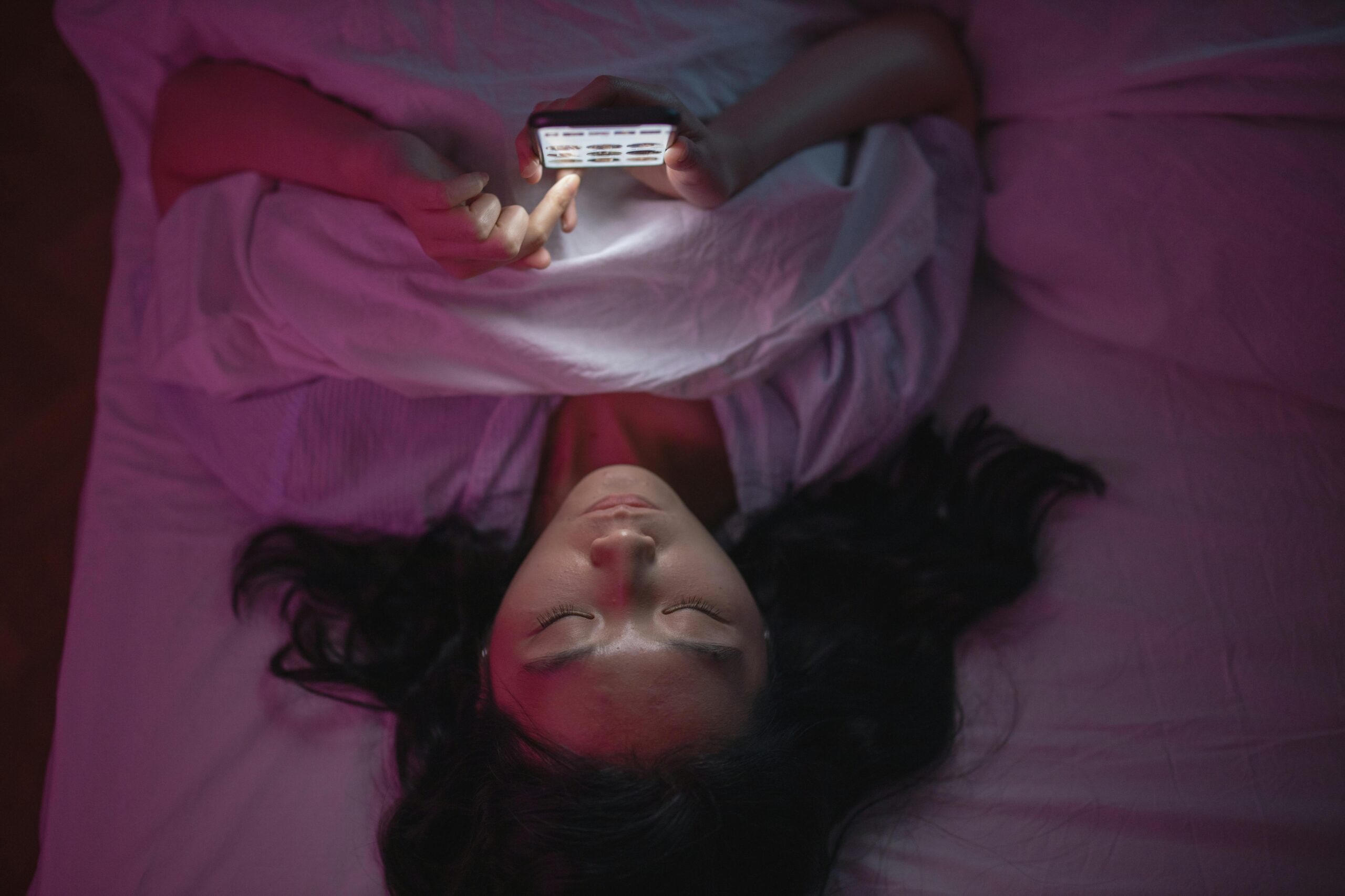 Young woman scrolling phone in bed with lights out seeking dopamine