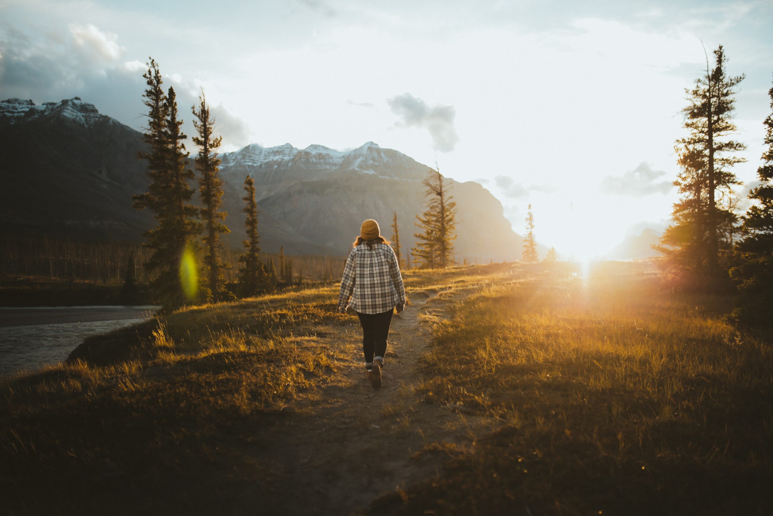 Person walking on a trail in the mountains with sun setting, in flannel, jeans and hat.