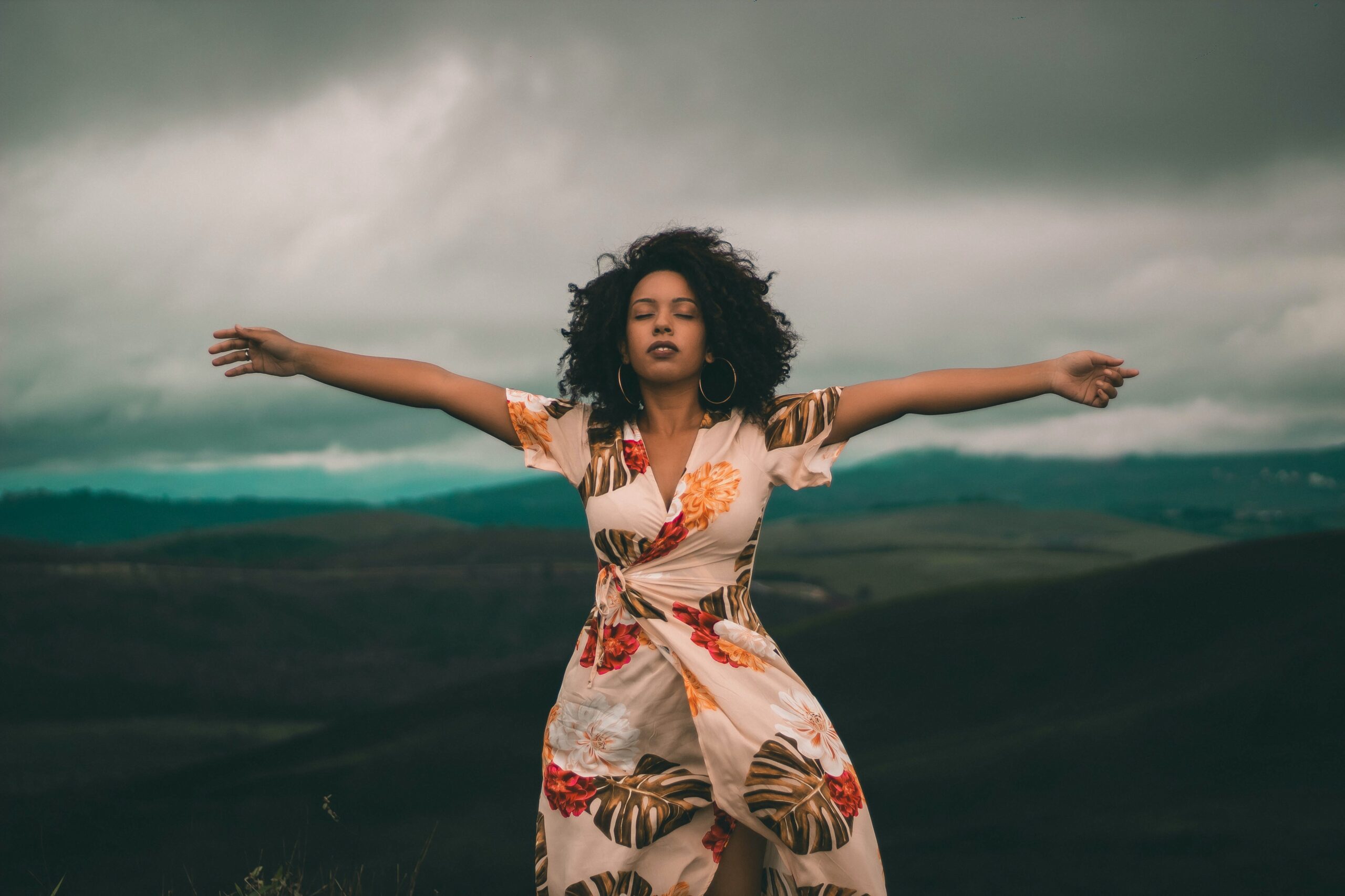 Image of a Black woman expressing a feeling of relief with arms spread wide, eyes closed, in floral dress in the mountains.