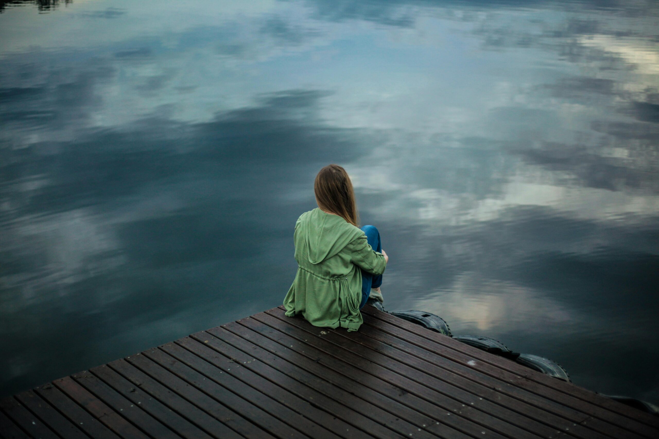 Person contemplating Ketamine Assisted Psychotherapy (KAP) to treat their depression, sitting on a dock over looking water with overcast sky