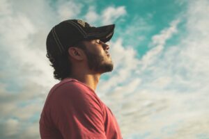 Young male with hat on closing eyes, head tilted up toward light. Blue sky in background with clouds. Practicing mindfulness