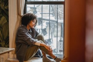 Young Black female sitting against a window, looking sad.