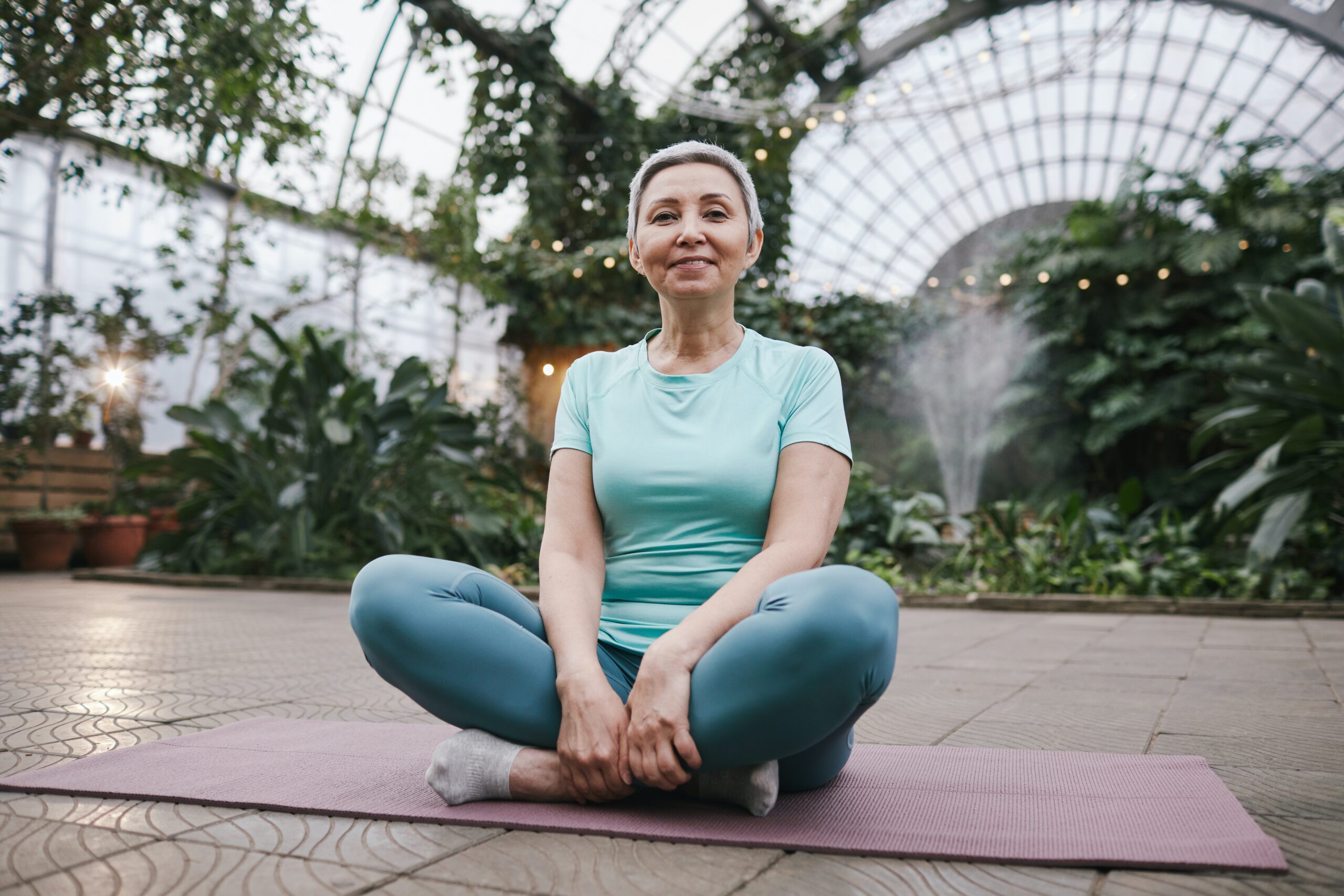 Middle-aged woman with short, white hair, sitting on a yoga mat in a atrium with blue yoga pants and blue shirt.