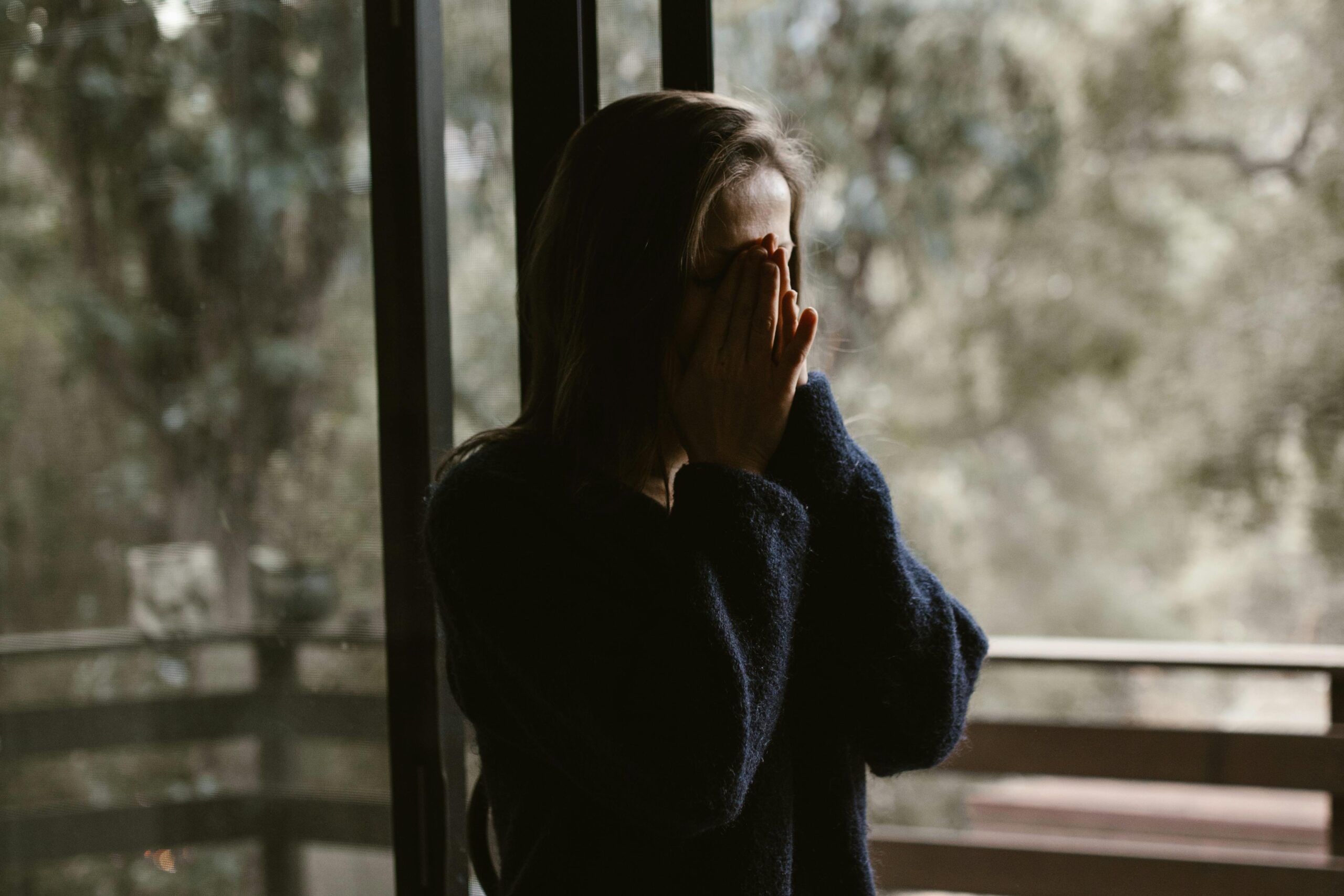 Person with chronic pain covering face next to a window