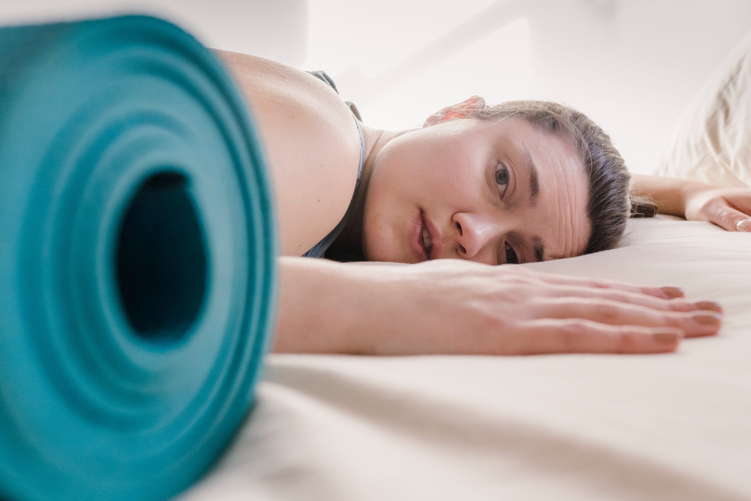 person lying exhausted on bed next to blue rolled-up yoga mat.